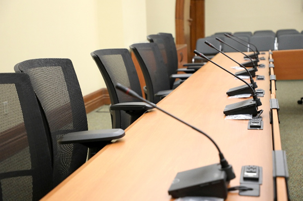 A row of empty chairs lined up along a desk with corresponding microphones in a committee meeting room.