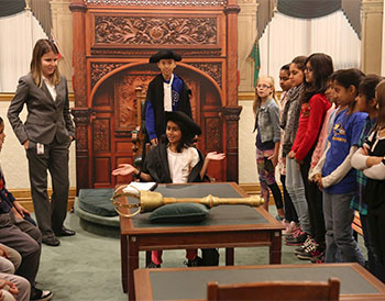 Group of young students surrounding a table with the Mace on top. Two students are in Parliamentary uniform acting as the Speaker and the Clerk. 